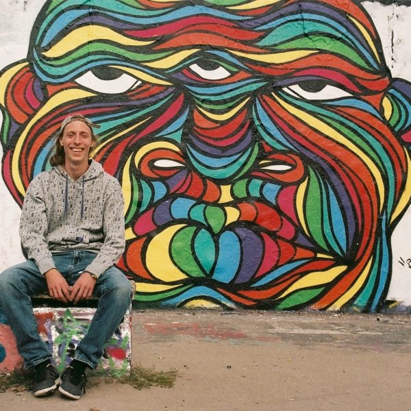 Hans From Space mit "Bruddha", Profilfoto mit Wandgestaltung / Graffiti in Regenbogen-Farben im Hintergrund, Berlin Mauerpark